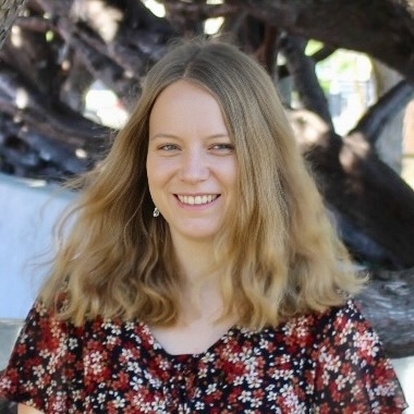 Portrait of a young blonde woman, smiling at the camera, standing in front of a tree.