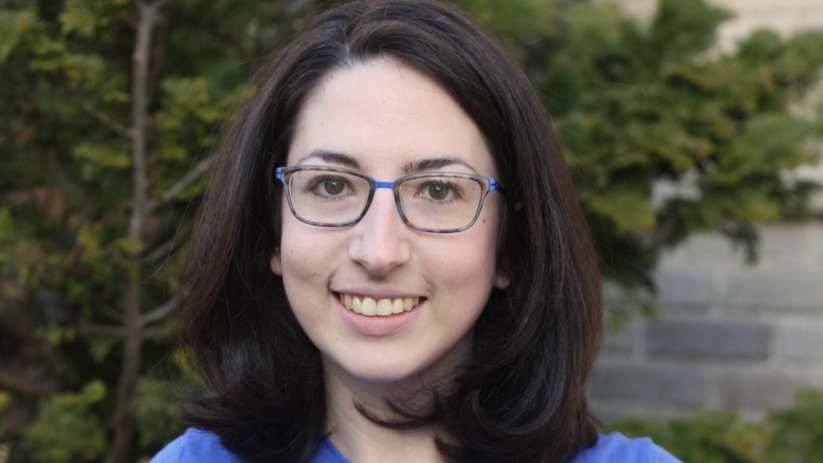 Close portrait of a young woman with dark hair and glasses, smiling.