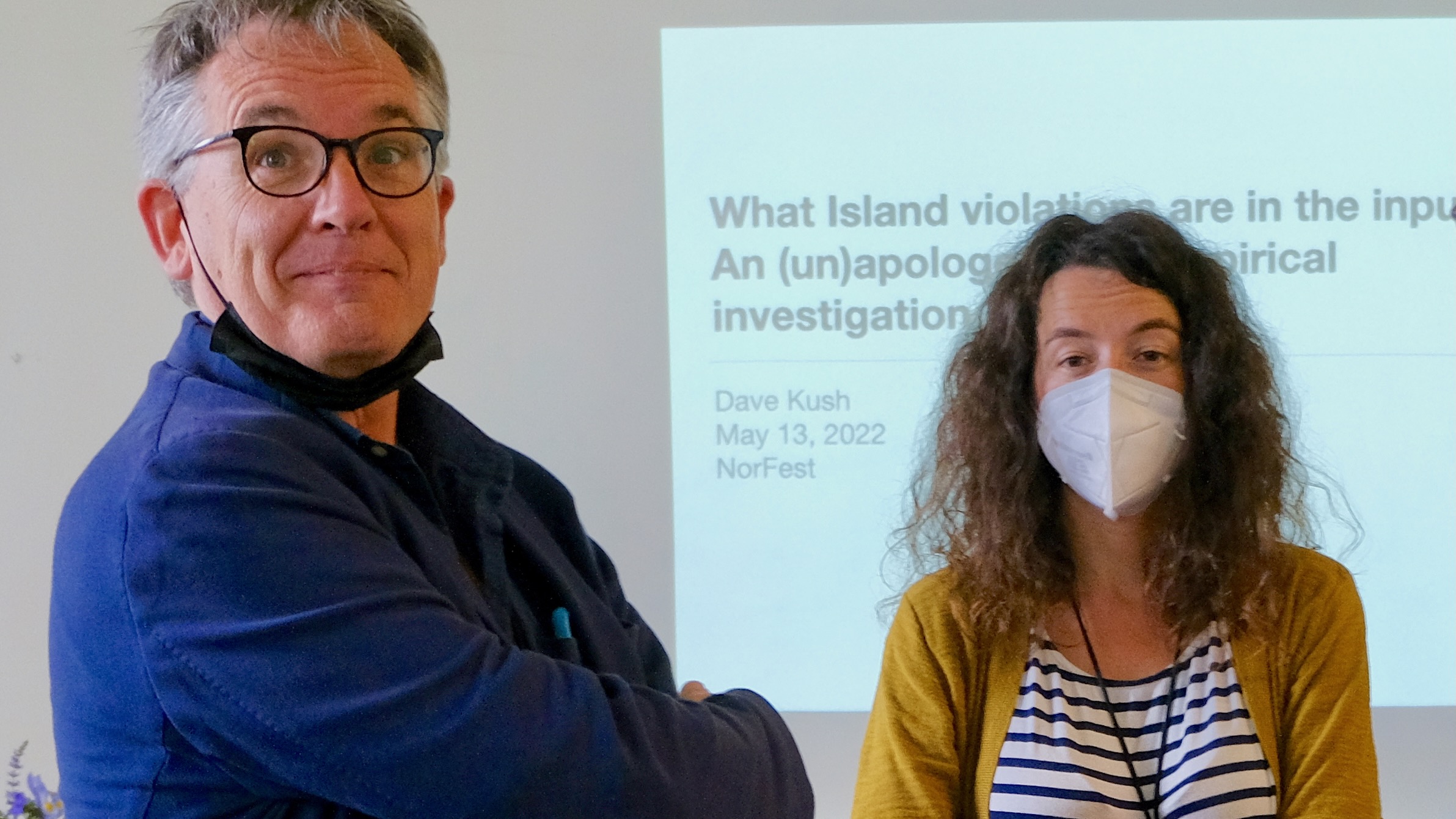 A man in a blue shirt staring whimsically at the camera, next to a more reserved woman in a mustard-colored cardigan and a COVID mask, in front of a screen showing a projected lecture slide.