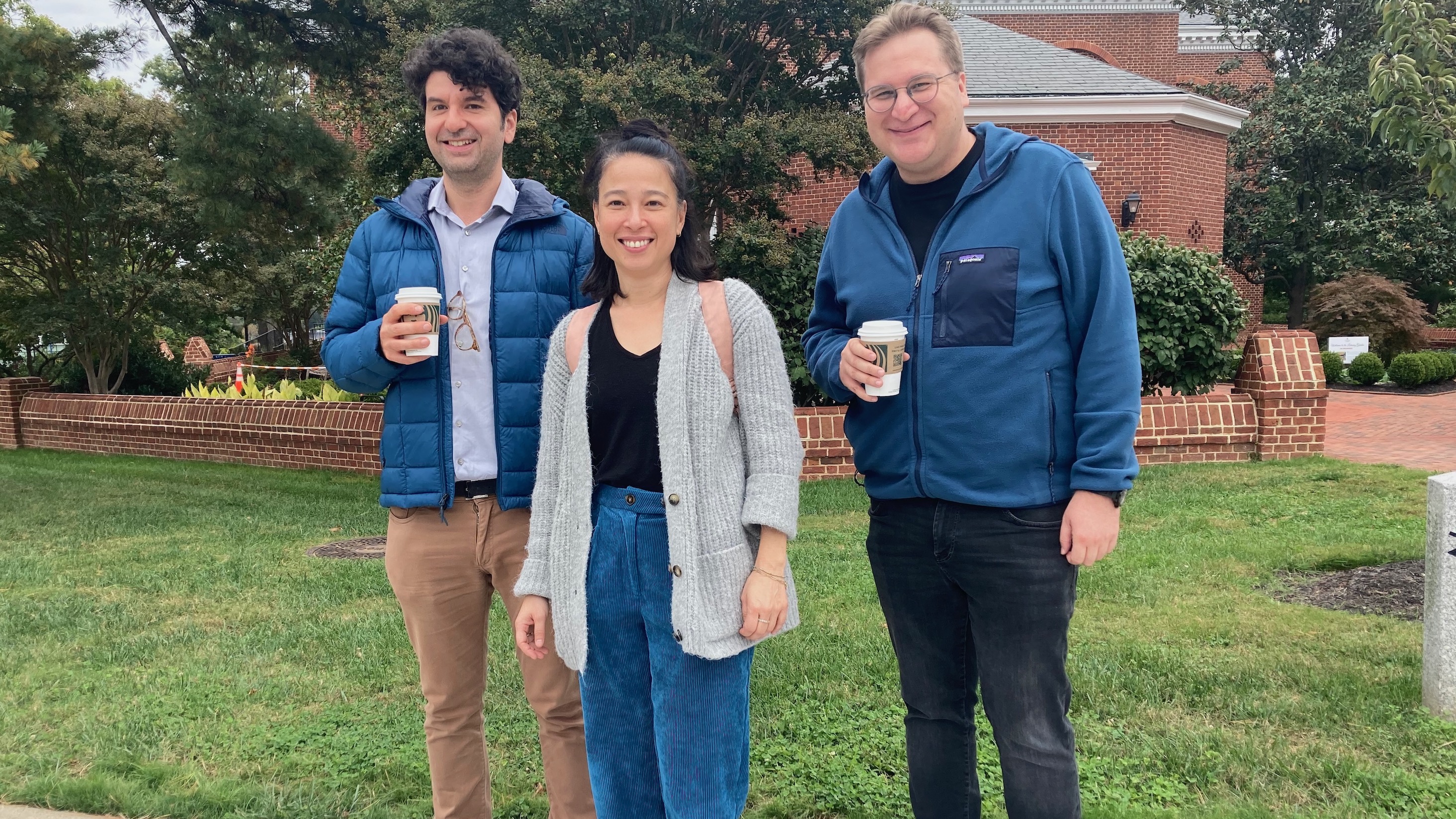 Two men and a woman standing outside posing, the two men in blue fleeces and holding coffee cups, flanking a woman wearing corduroy pants in exactly the same shade of blue.