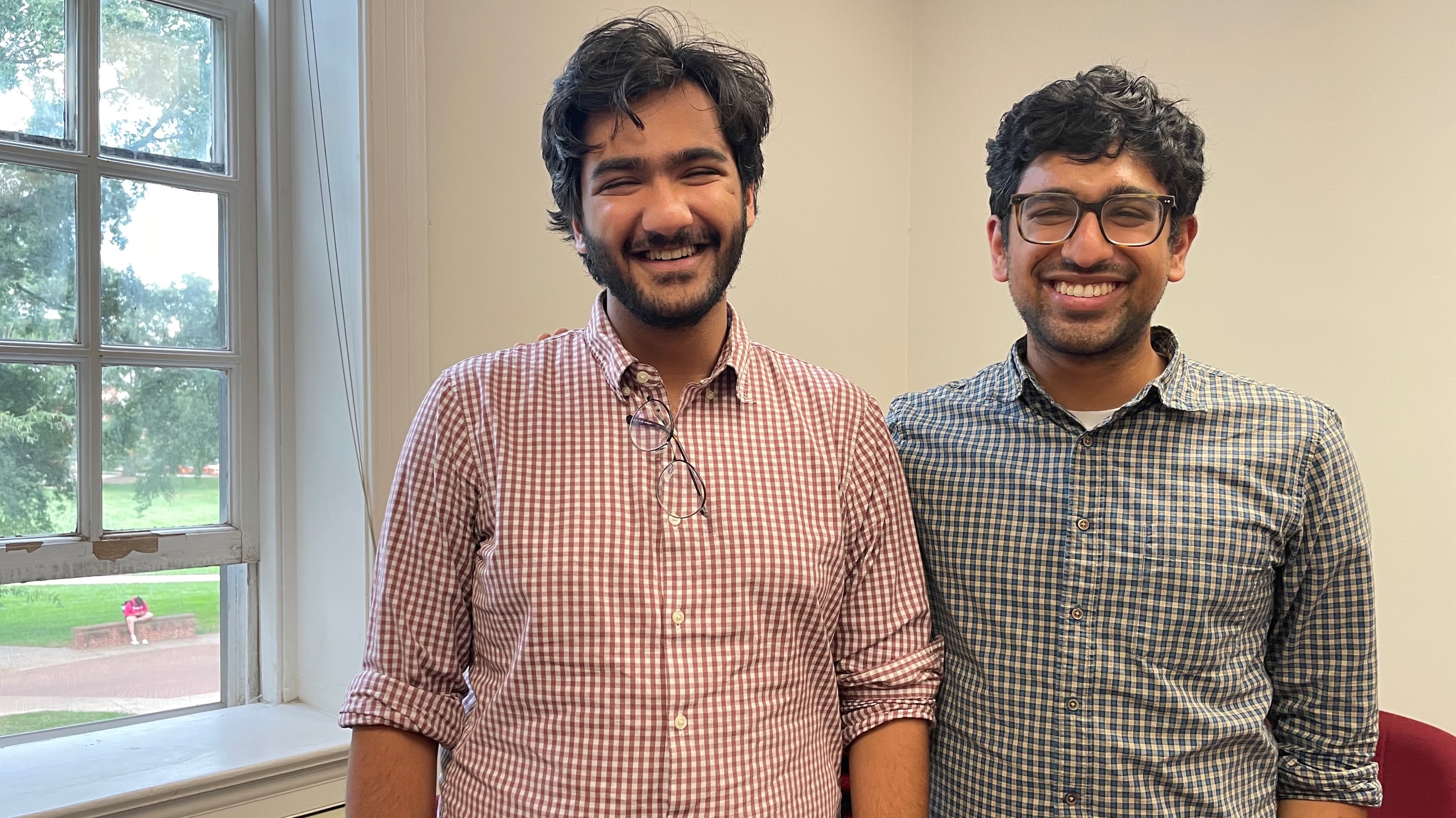 Two young men standing next to each other, one in a red checked shirt, the other in a grey checked shirt.