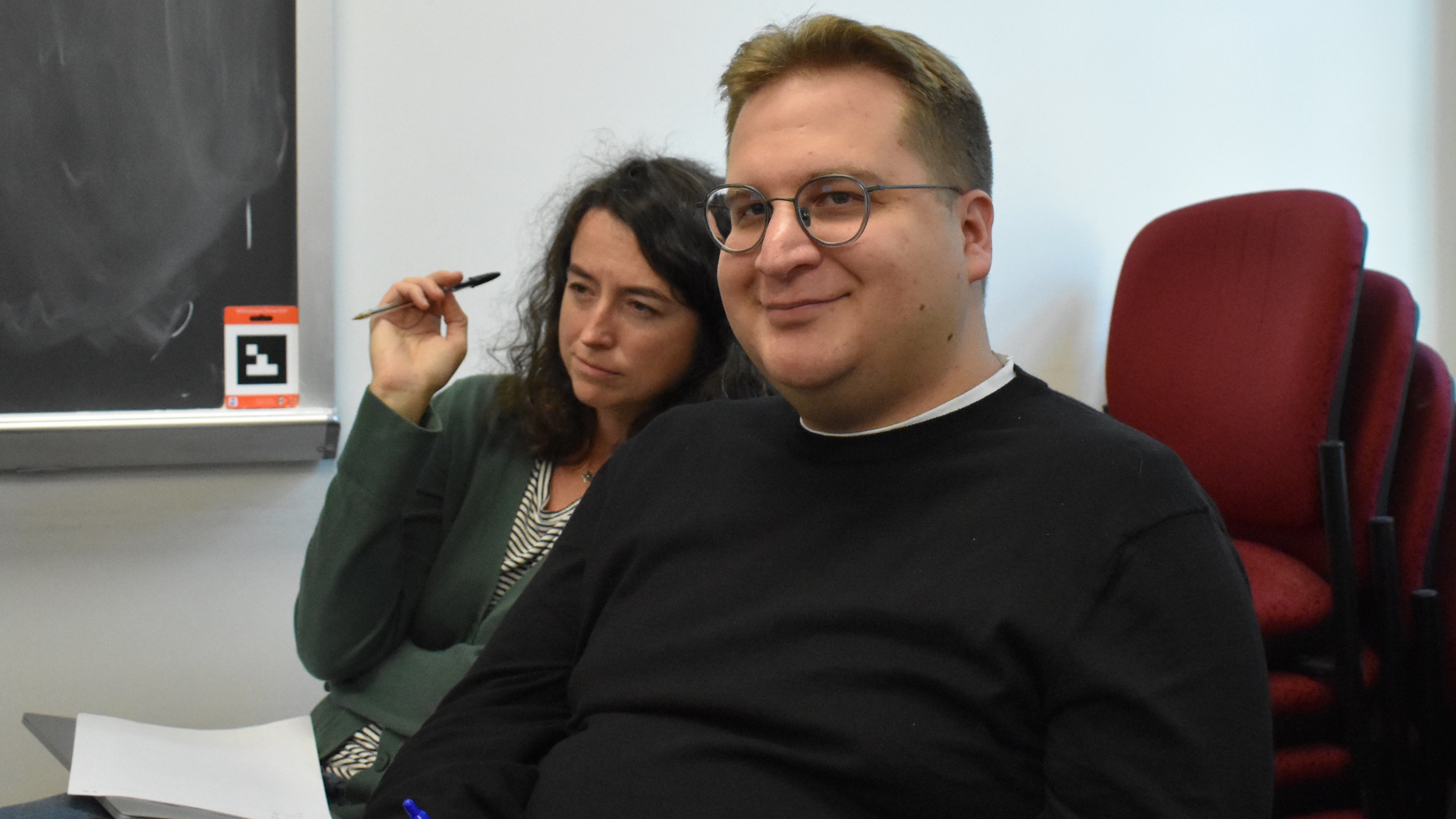 A young man with glasses, sitting in a classroom, with a woman behind him, looking thoughtful.