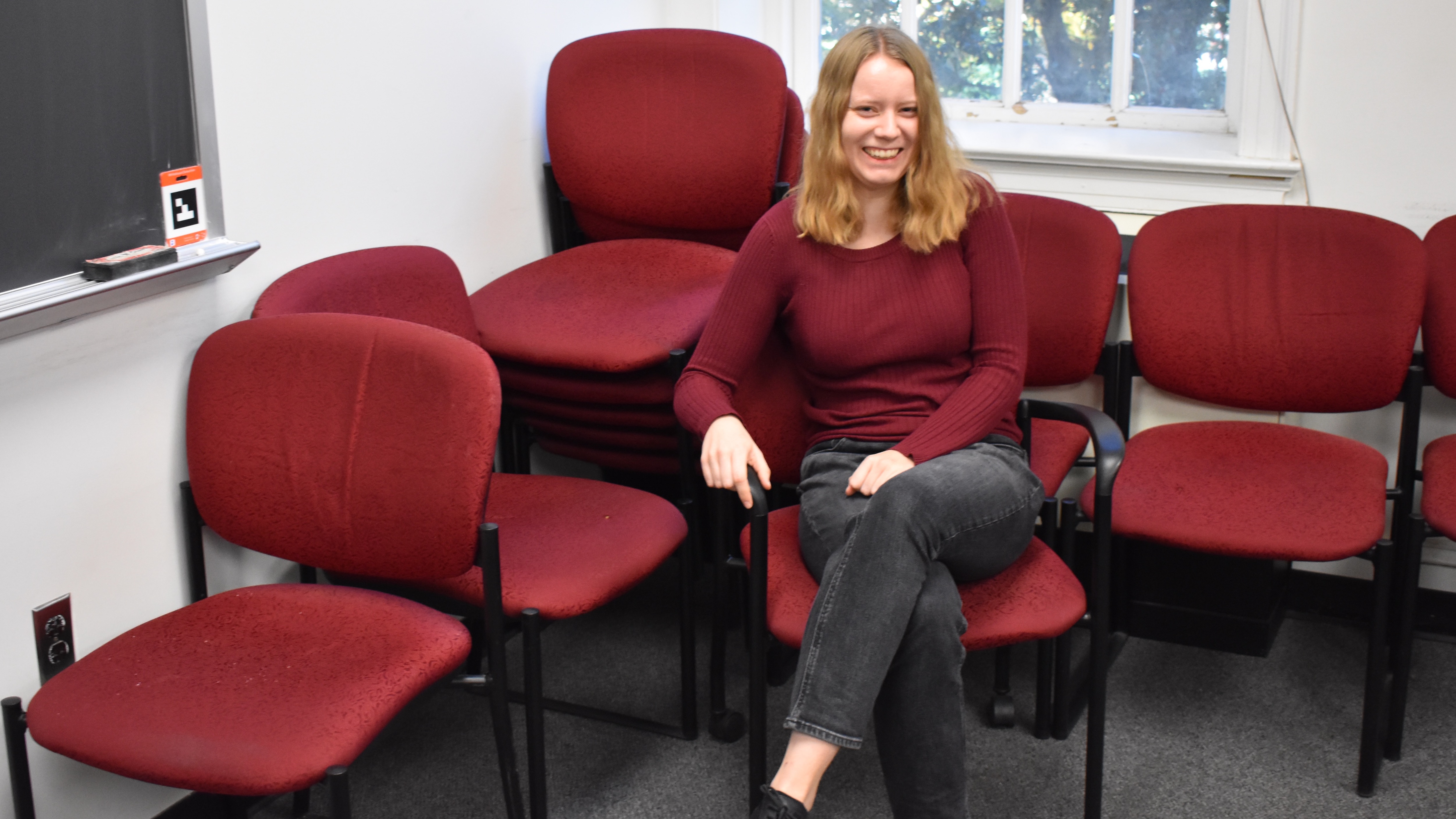 A young woman, wearing a burgundy pullover and charcoal jeans, sitting amidst an array of burgundy office chairs on a charcoal carpet.