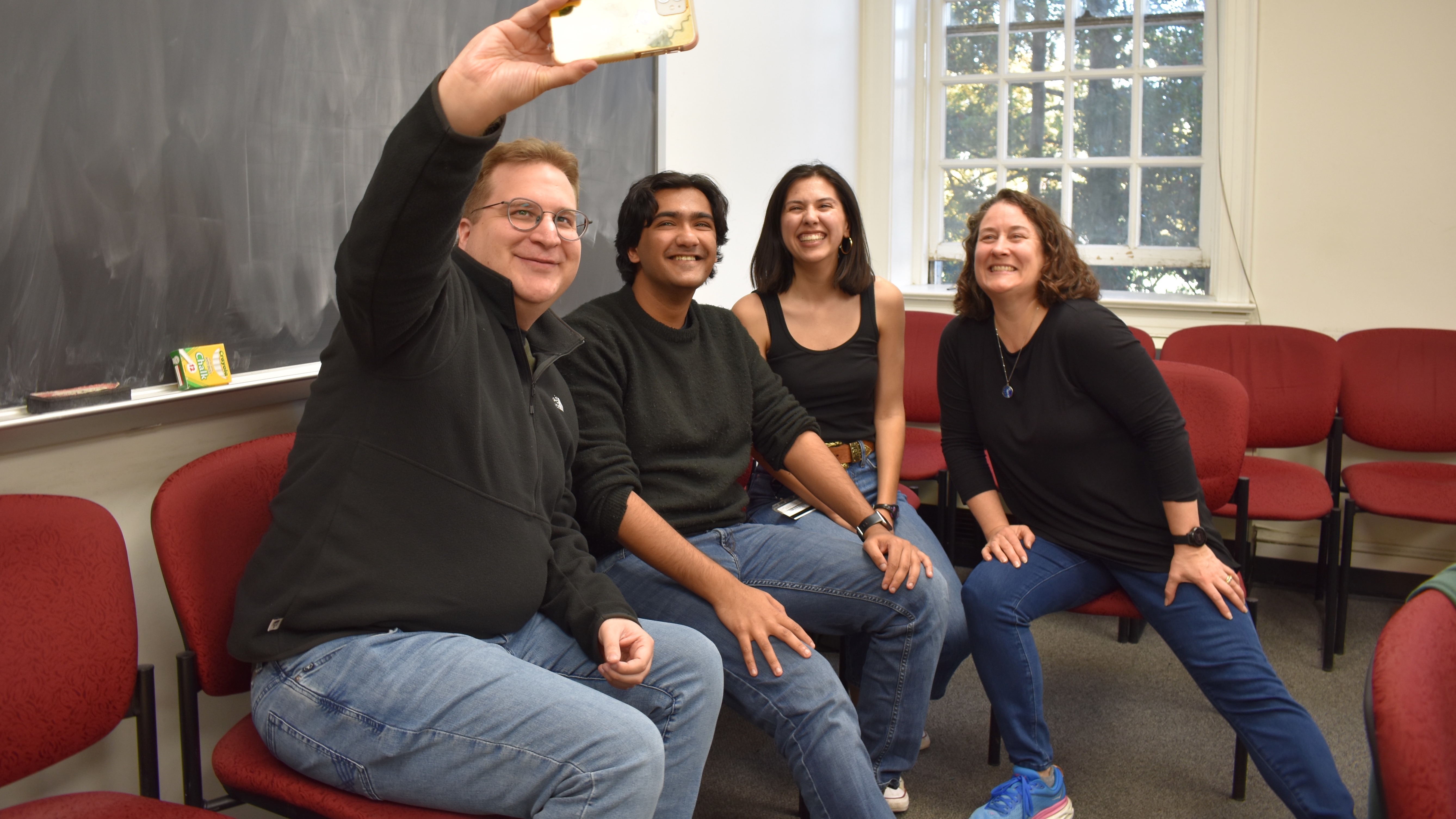 Four people all wearing blue jeans and a black top, sitting in a classroom, taking a selfie of themeselves.