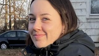 Close portrait of a young woman in profile, outside in seemingly cold weather, a car in a driveway visible in the background.