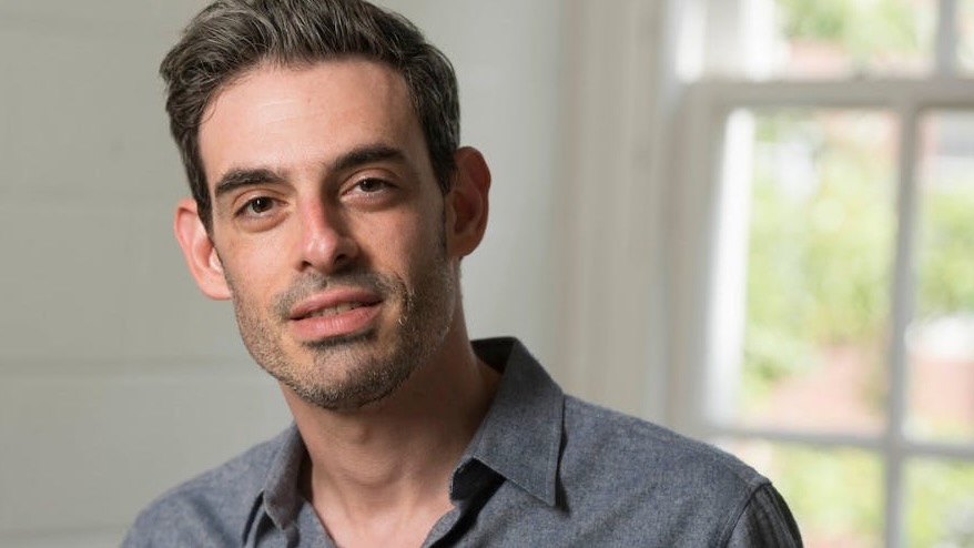 Close portrait of a man in a well-lit room, grinning confidently.