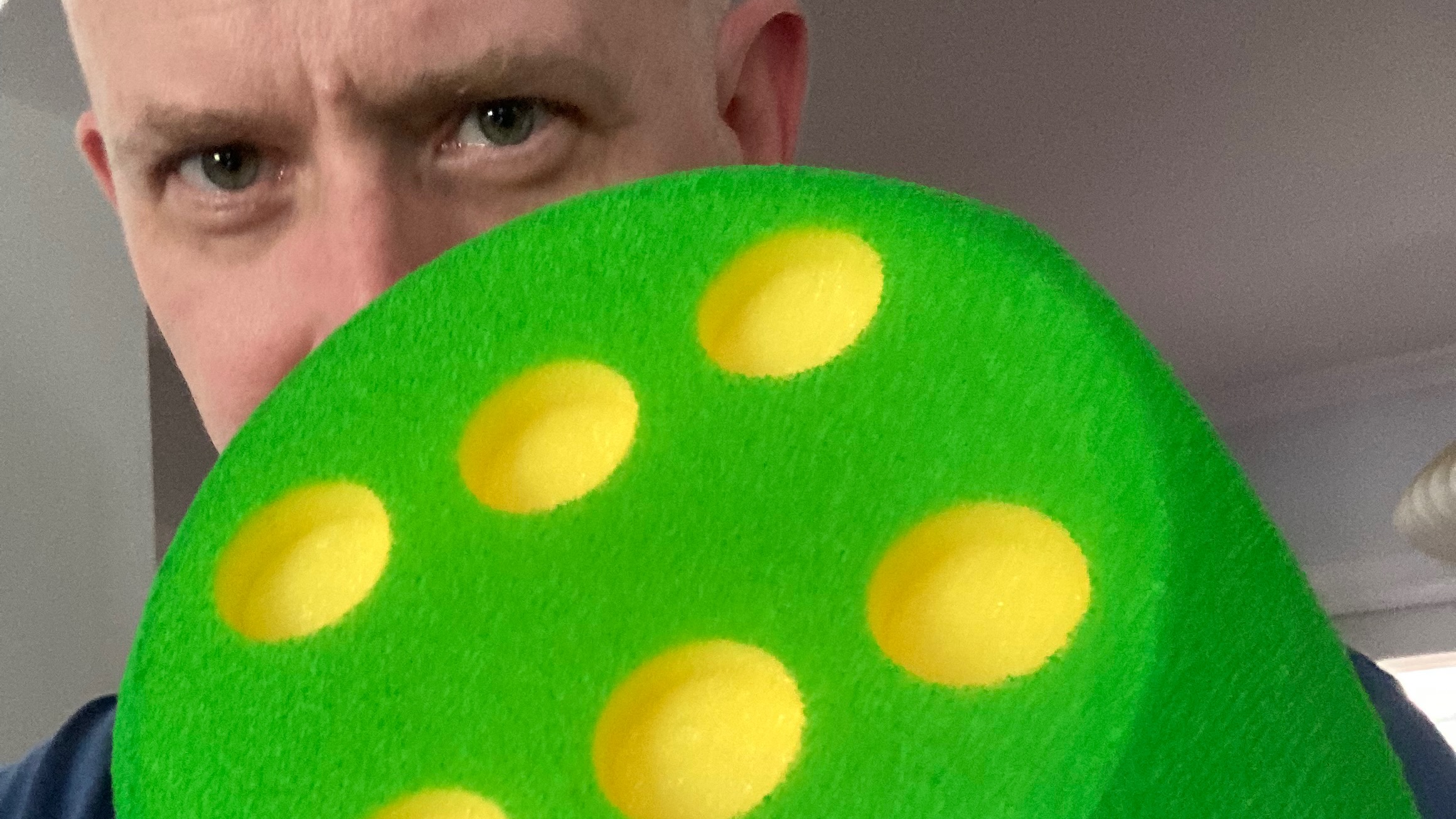 A close portrait of a very large green foam die, the six face pointed at the camera, and the mysterious eyes of its holder staring from the background.