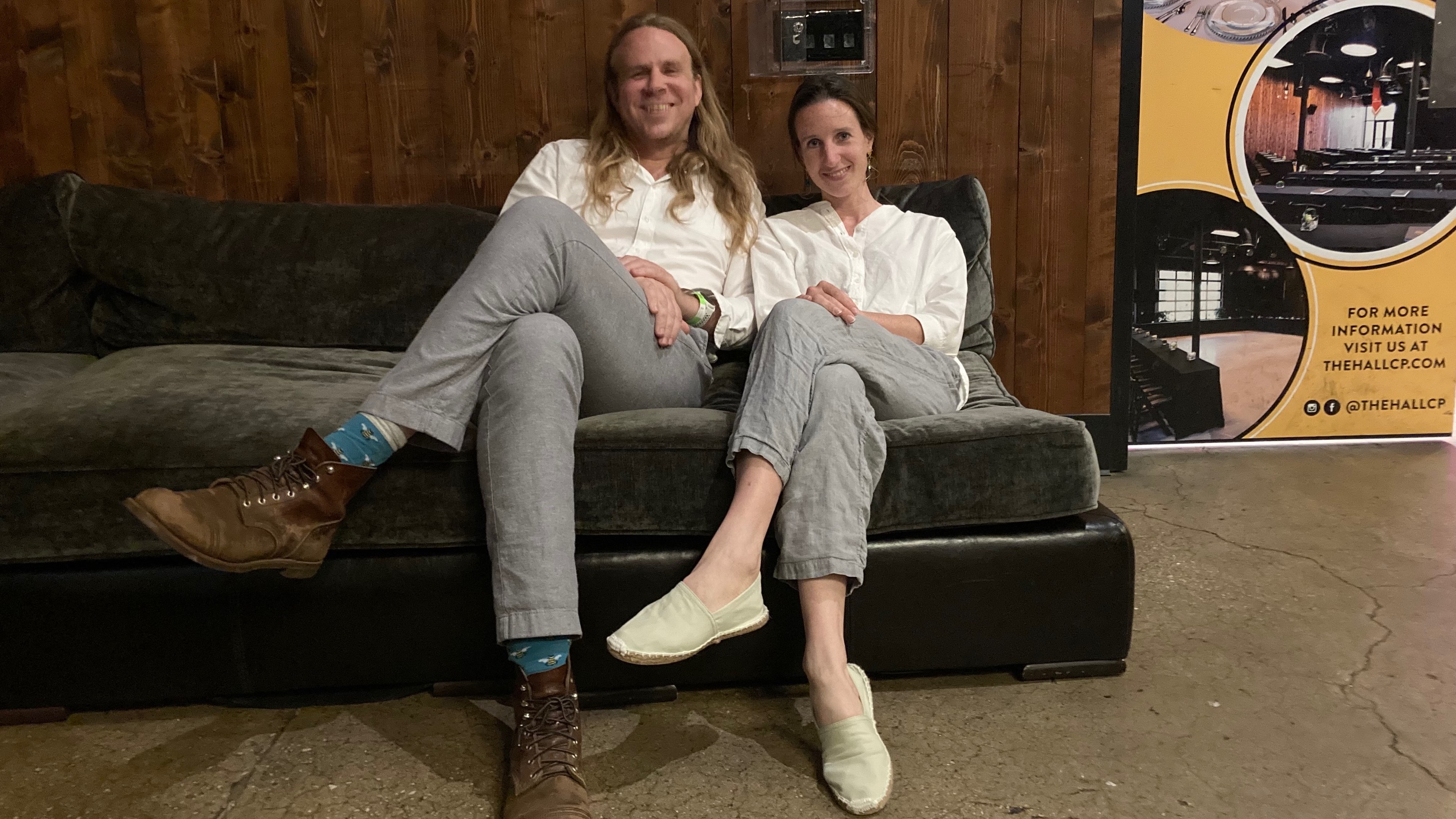 A young man and a young woman, seated cross legged on a brown leather sofa, both wearing seersucker pants and a white linen shirt.