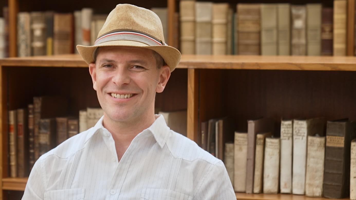 A man in a trilby hat, smiling, sitting in front of a bookcase.