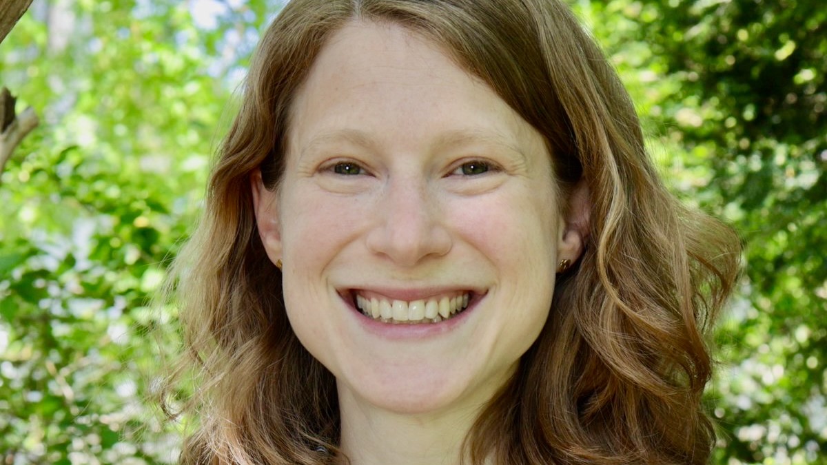 Close portrait of the face of a young woman smiling, standing outside against a background of tree leaves.