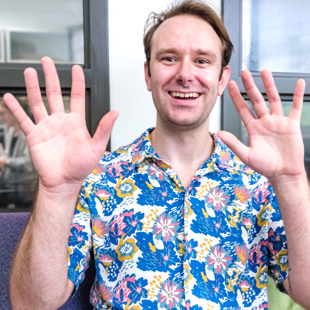 A young man, in a polynesian floral print shirt, holding up both palms facing the camera, smiling.