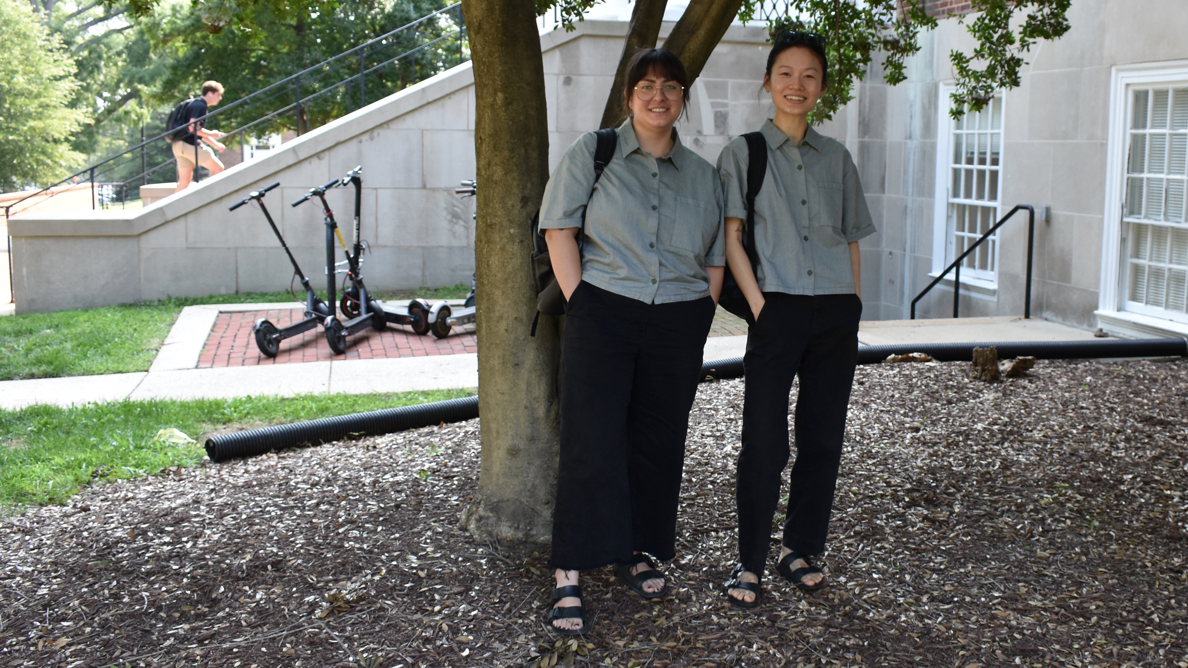 Two young women wearing exactly the same sage and black outfit, standing under a tree. 