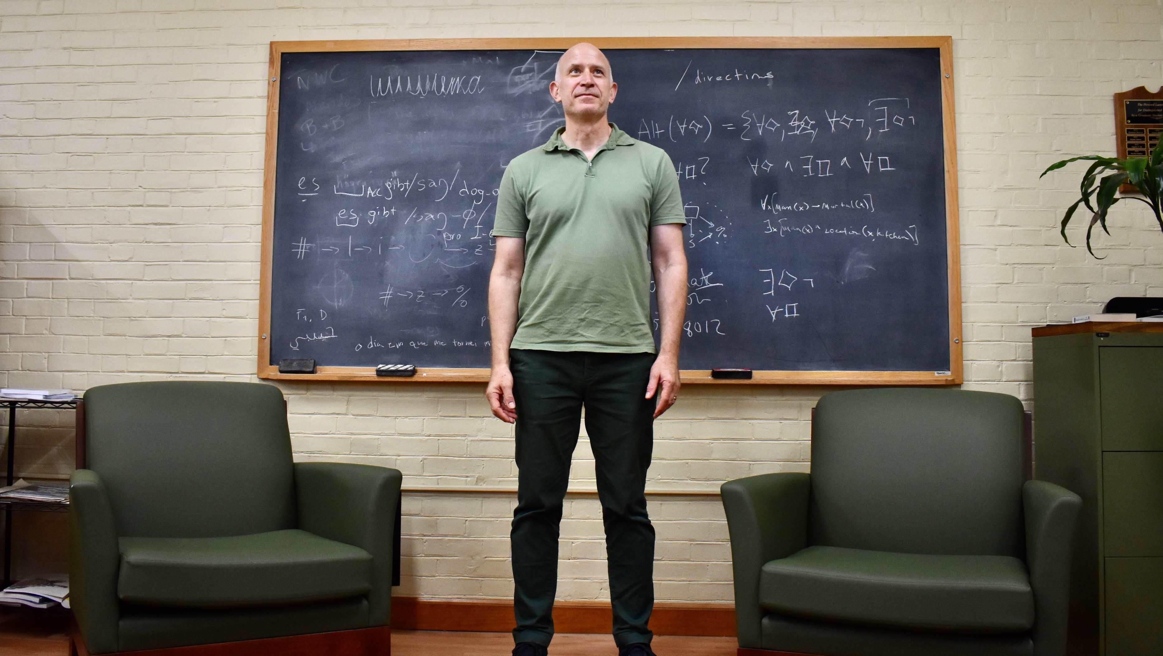 A man in dark green pants and a lighter green shirt standing between two dark green sofa chairs in front of a university blackboard.