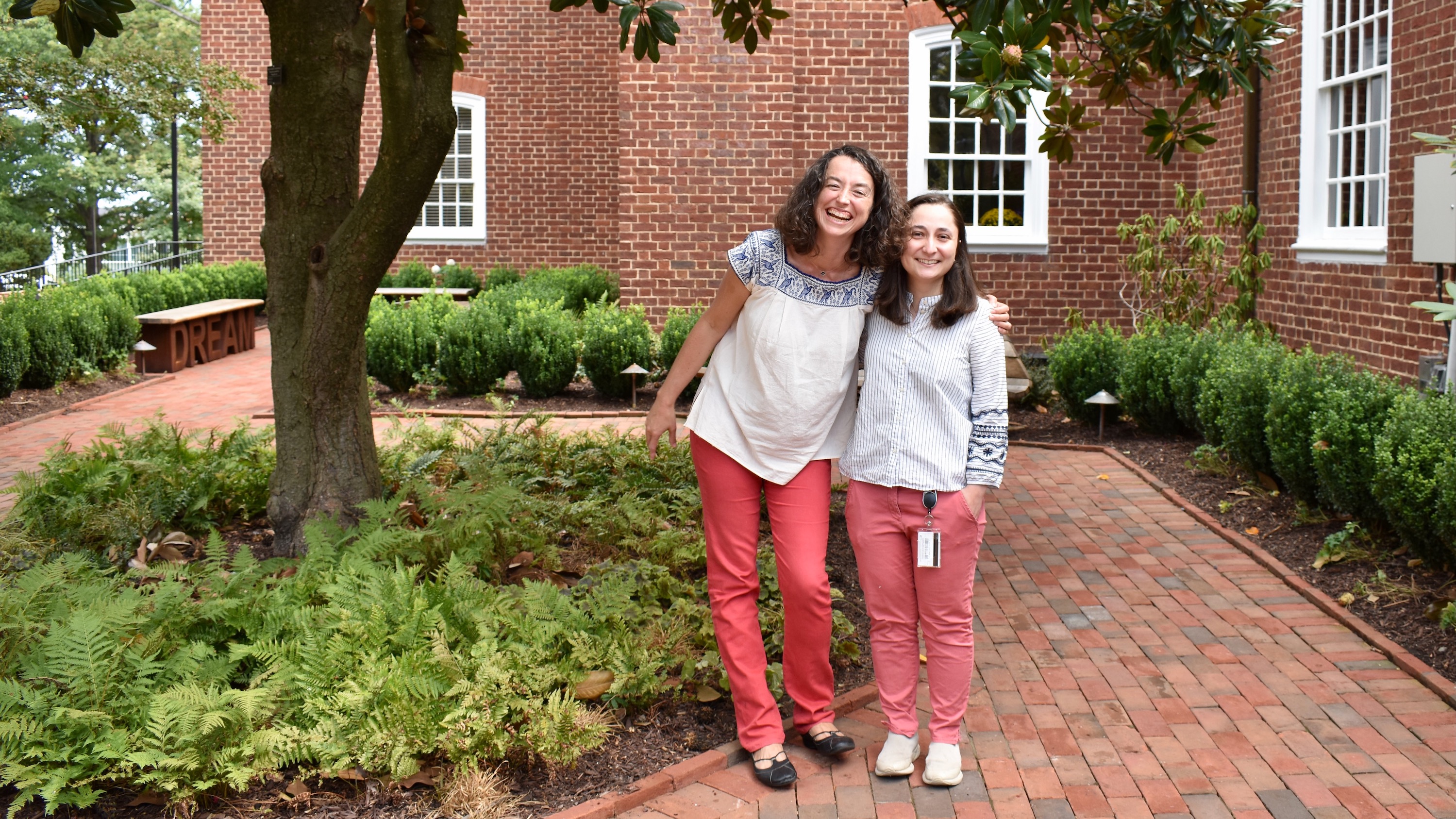 Two women standing in a garden, both wearing coral pants and embroidered tunics.