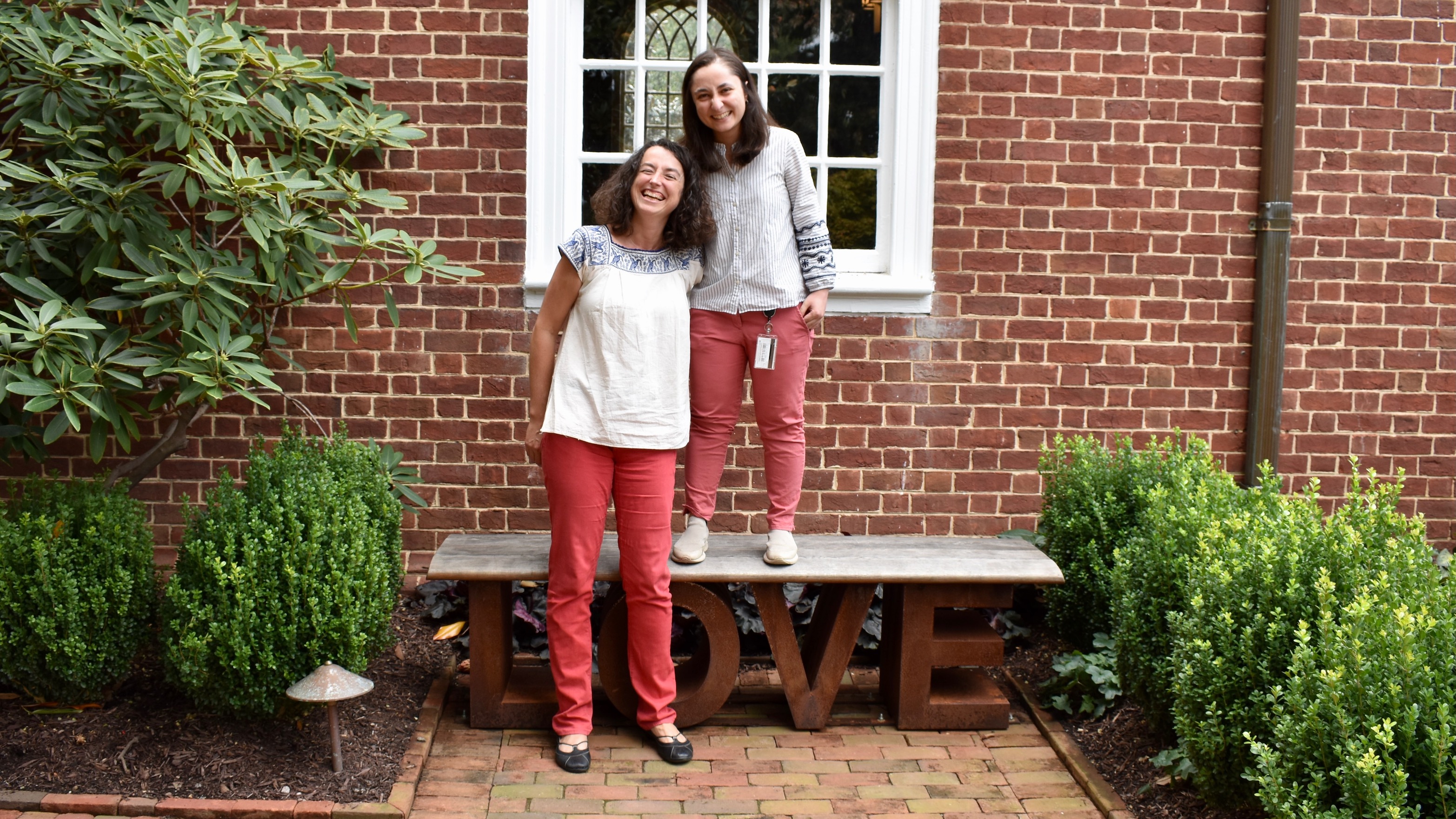 Two women standing in a garden, the shorter of the two standing on top of a bench, which is built in the shape of the word "love".