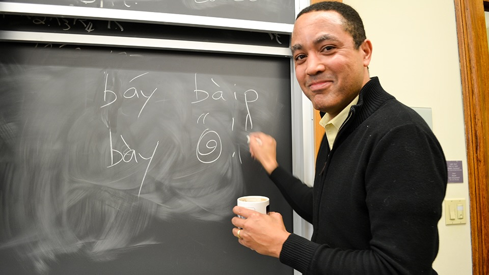 A man writing on a chalkboard, smiling back at the camera, holding a paper cup of coffee in his left hand.