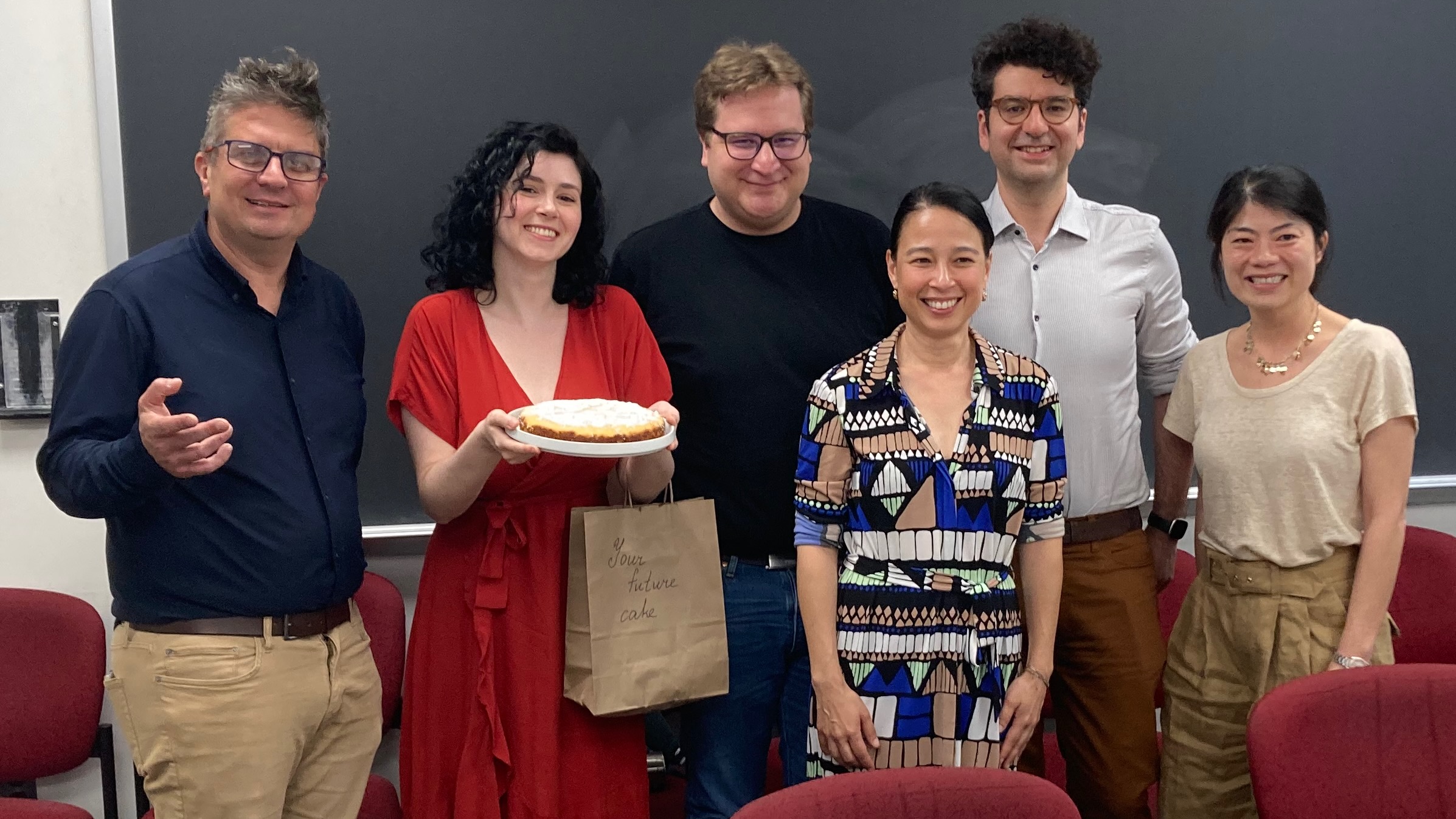 A group of six people standing together in front of a blackboard, one holding a cake, celebrating a dissertation defense.