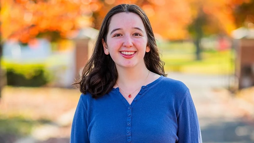 A young woman standing outside in front of bright orange fall foliage, wearing a royal blue shirt and smiling.