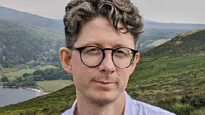 A man wearing glasses, standing on a how above a Scottish loch.