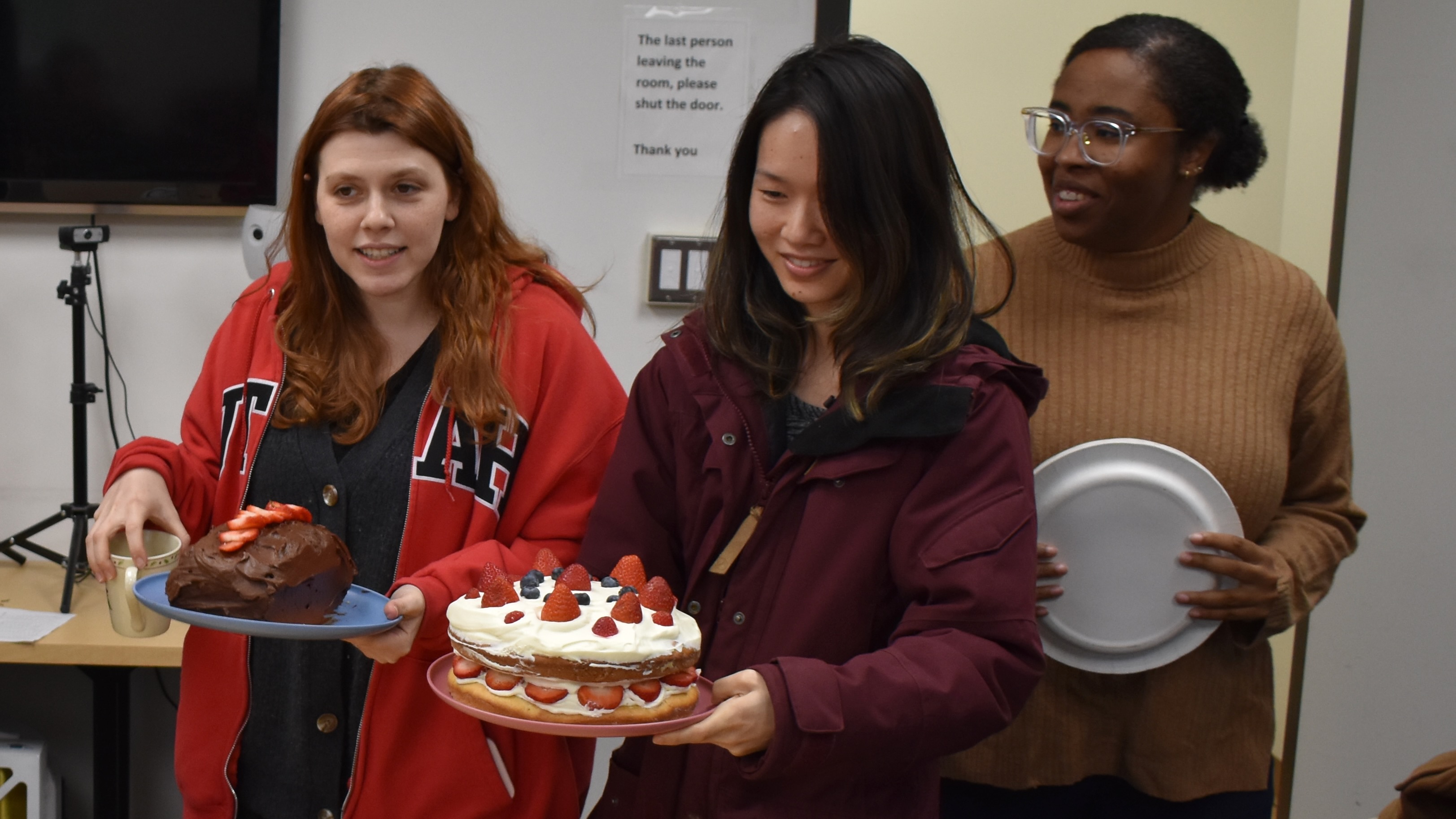 Three young women bringing two cakes and some plates into a room, evidently for a celebration.