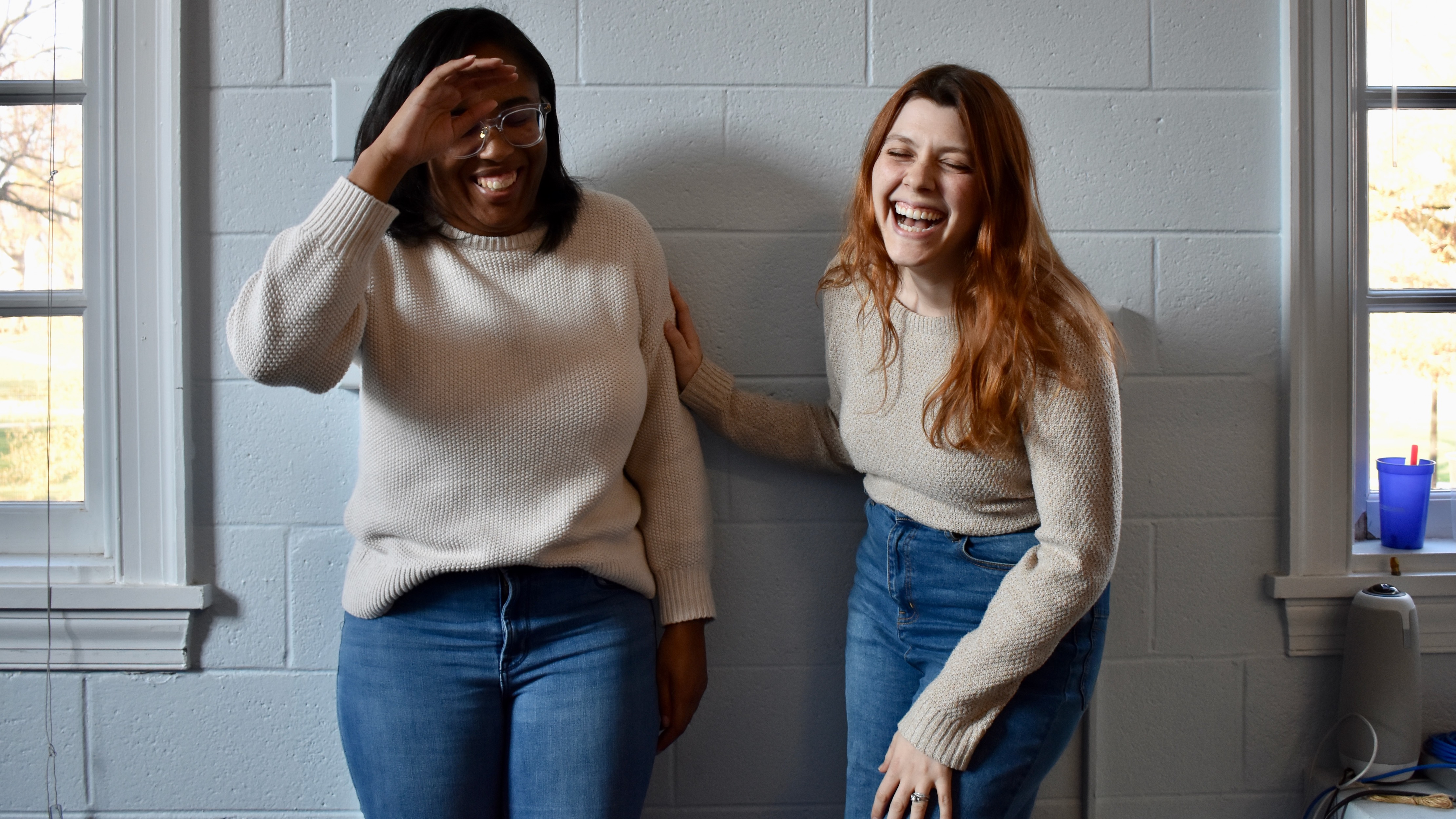 Two young women laughing, both wearing oatmeal colored cotton sweaters and blue jeans.