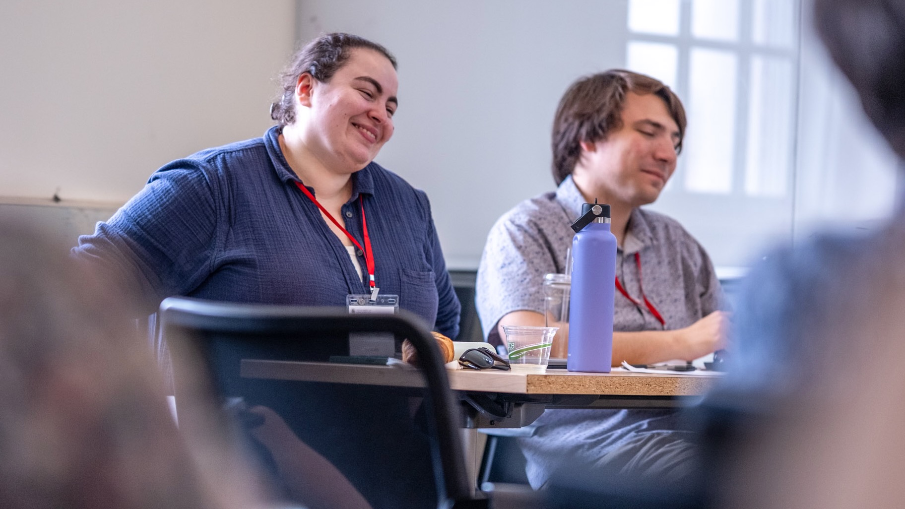 A young woman and a young man, sitting at a workshop, laughing.
