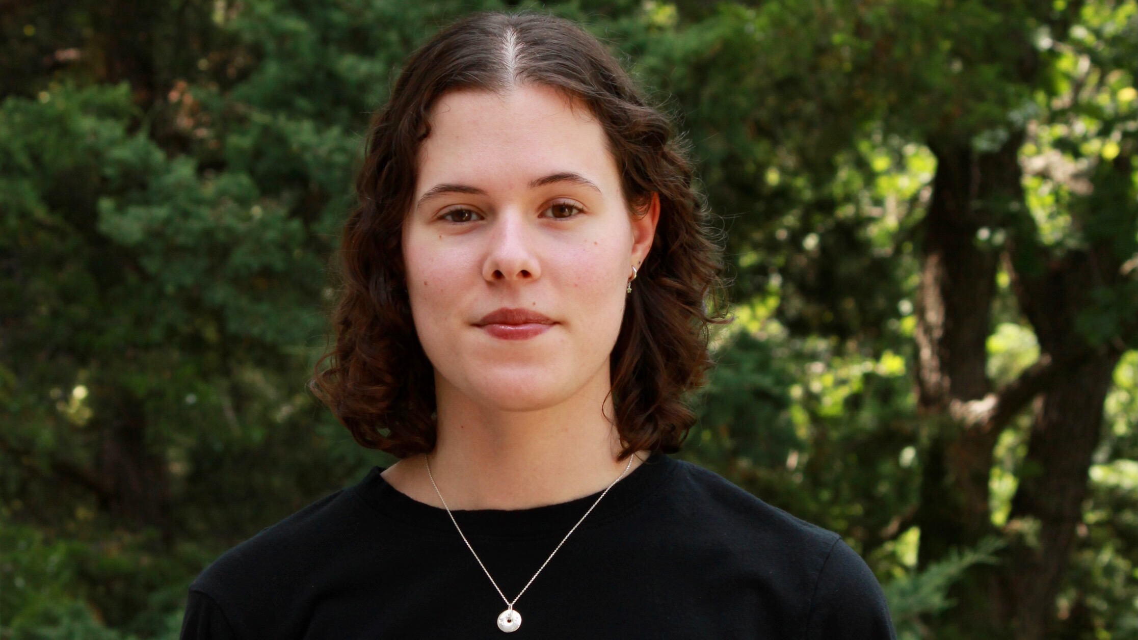 Portrait of a young woman in a black sweater, standing outside with dark trees behind her.