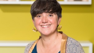 A woman smiling, sitting in front of a yellow wall.