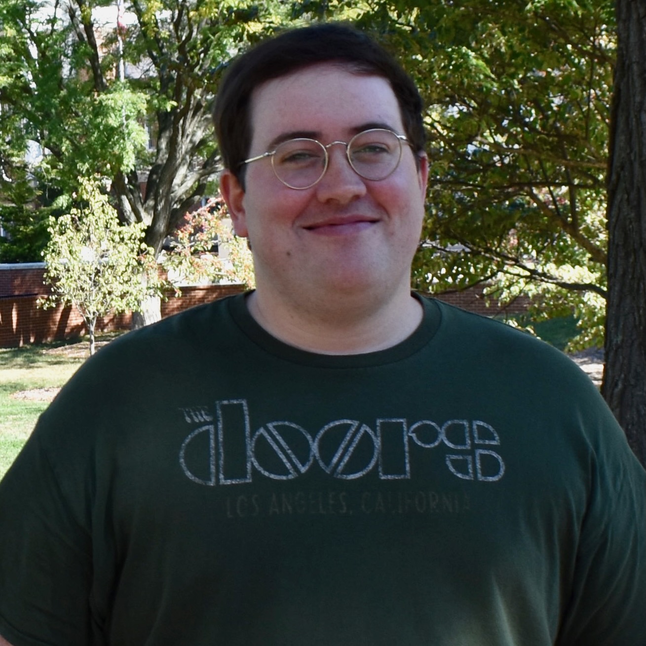 A young man in a green Doors t-shirt, standing outside in the shade on a sunny day, smiling for the camera.