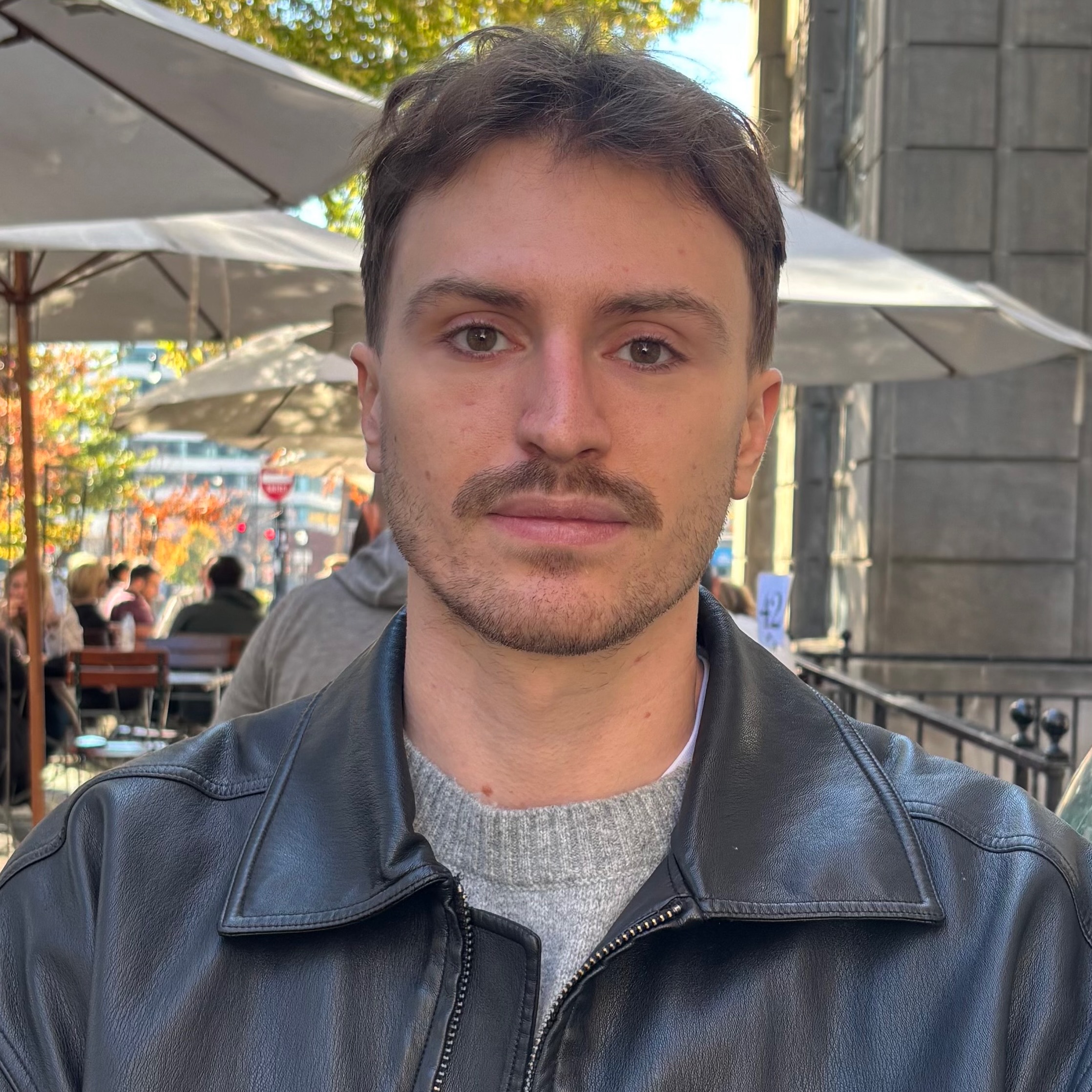 A close portrait of young man in a black leather jacket sitting outside at a sidewalk cafe, gazing gently at the camera.