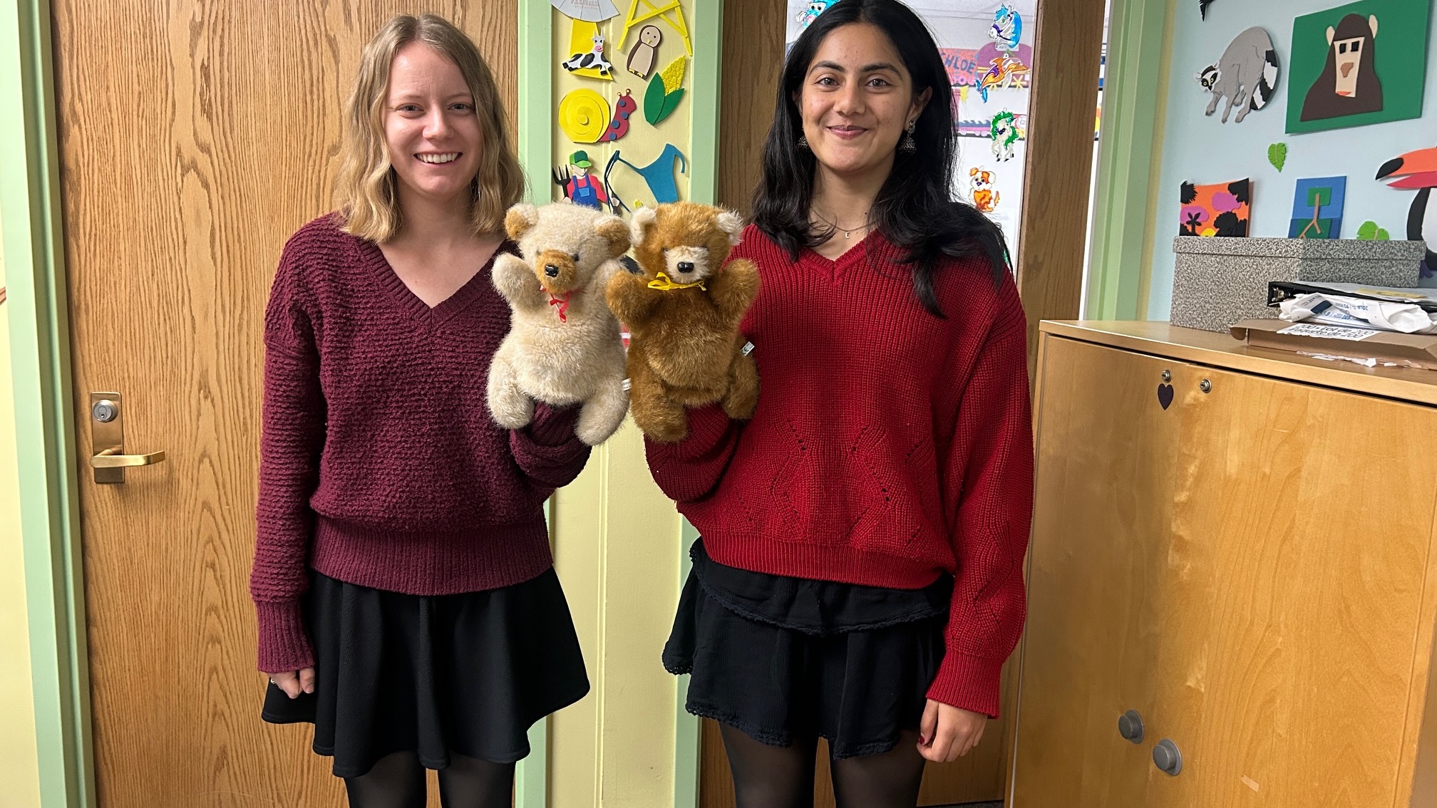 Two young women in nearly identical outfits, standing in mirror image poses, each holding up a stuffie and smiling at the camera.