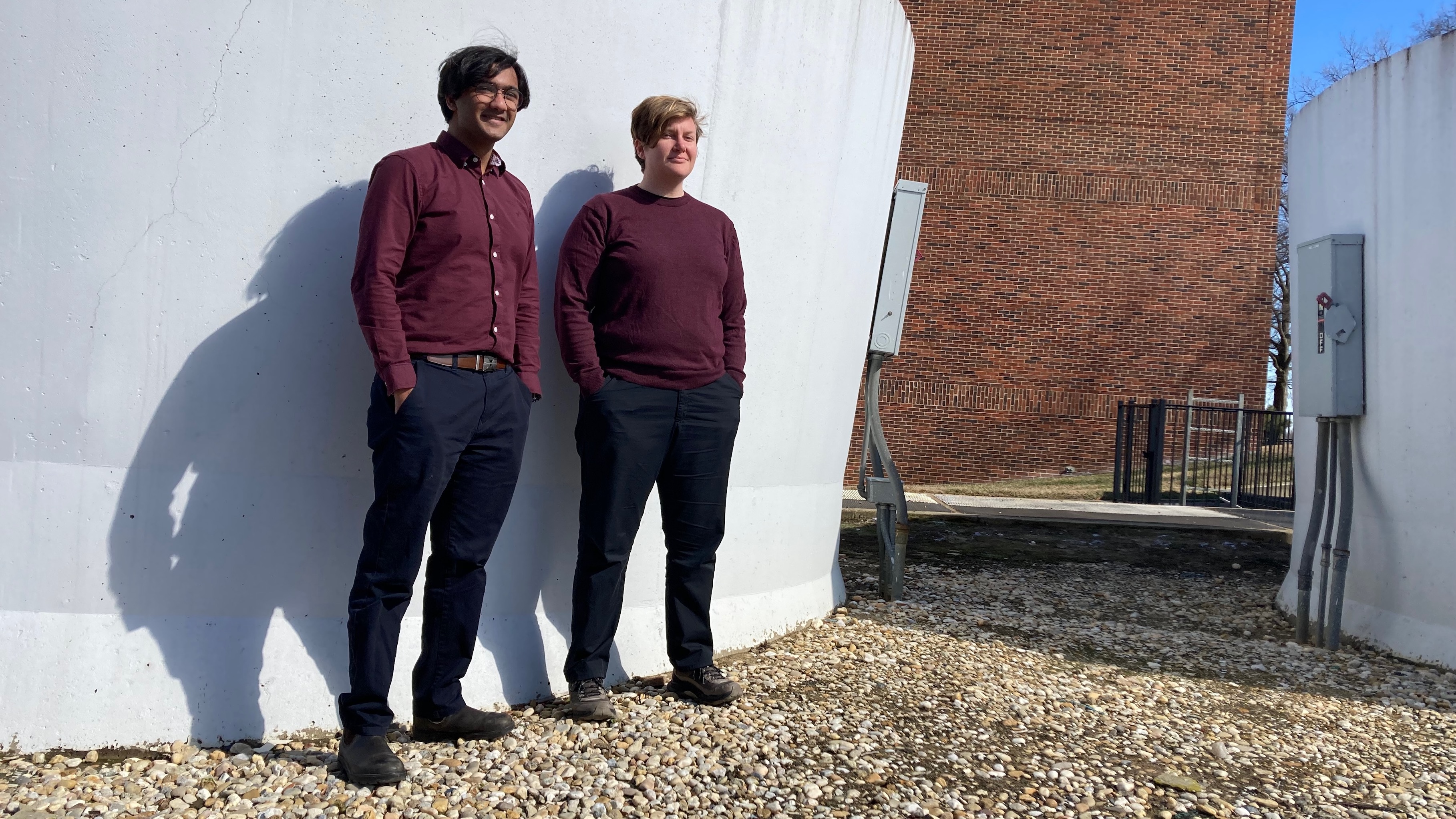 A young man and young woman, both in burgundy tops and midnight blue pants, standing outside on a bright blue day, in front of a massive concrete HVAC chimney, painted white.