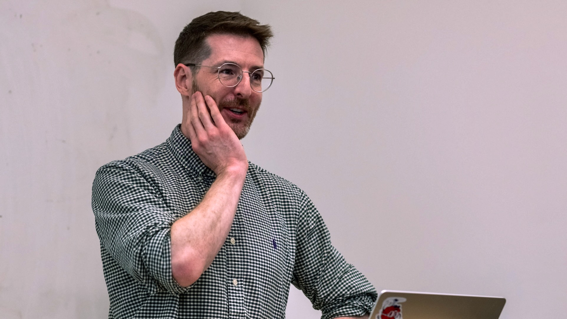 A man, standing at a lectern with his hand pensively on his cheek, smiling and listening.