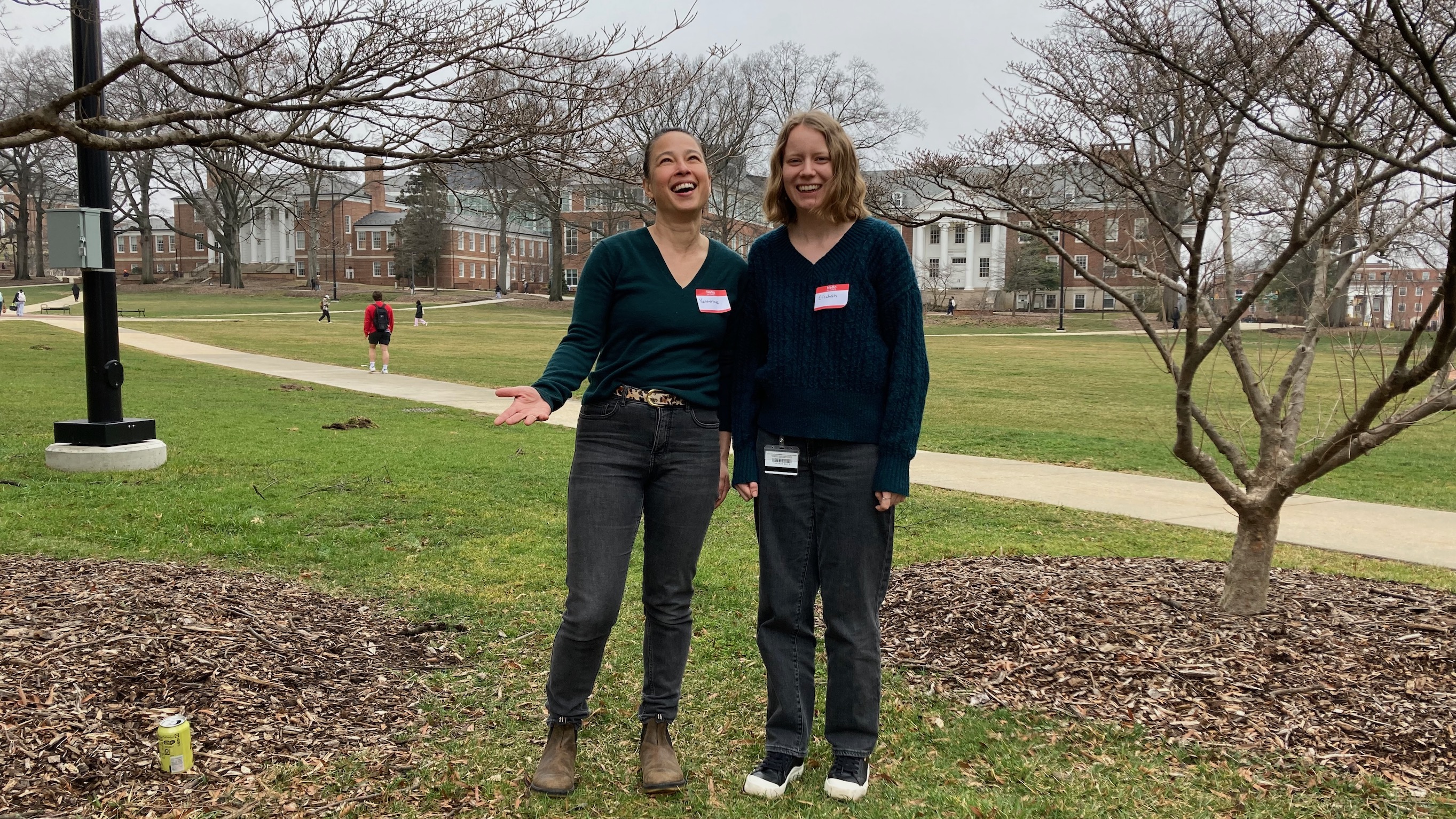 Two women standing outside in the early spring, both wearing dark teal sweaters and charcoal grey jeans. 