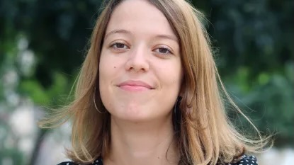 A young woman standing outside, chin up, grinning as her hair blows lightly in a slight breeze.