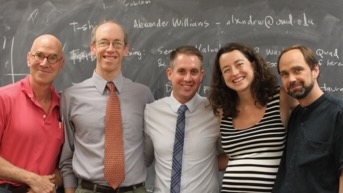Five people, standing close to each other in front of a chalkboard, smiling.
