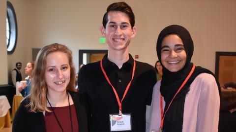 Three undergraduate students, two girls and a boy, arm in arm, smiling at the camera